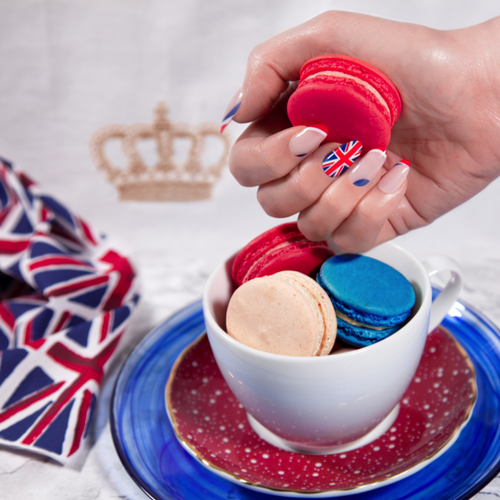 Hand with red nail polish holding a red macaron over a cup filled with colorful macarons on a blue plate with a Union Jack design.